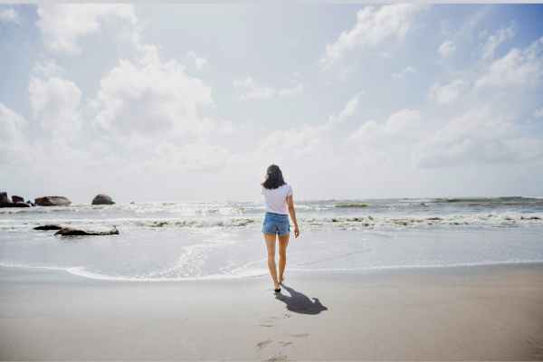woman walking along the shore of a beach wearing a Tshirt and shorts on blue sky day
