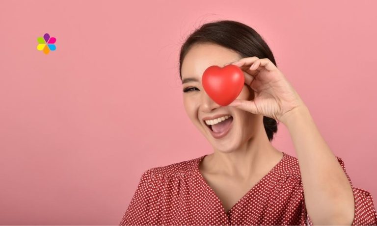 woman in red shirt holds red heart shape to her eye and smiles