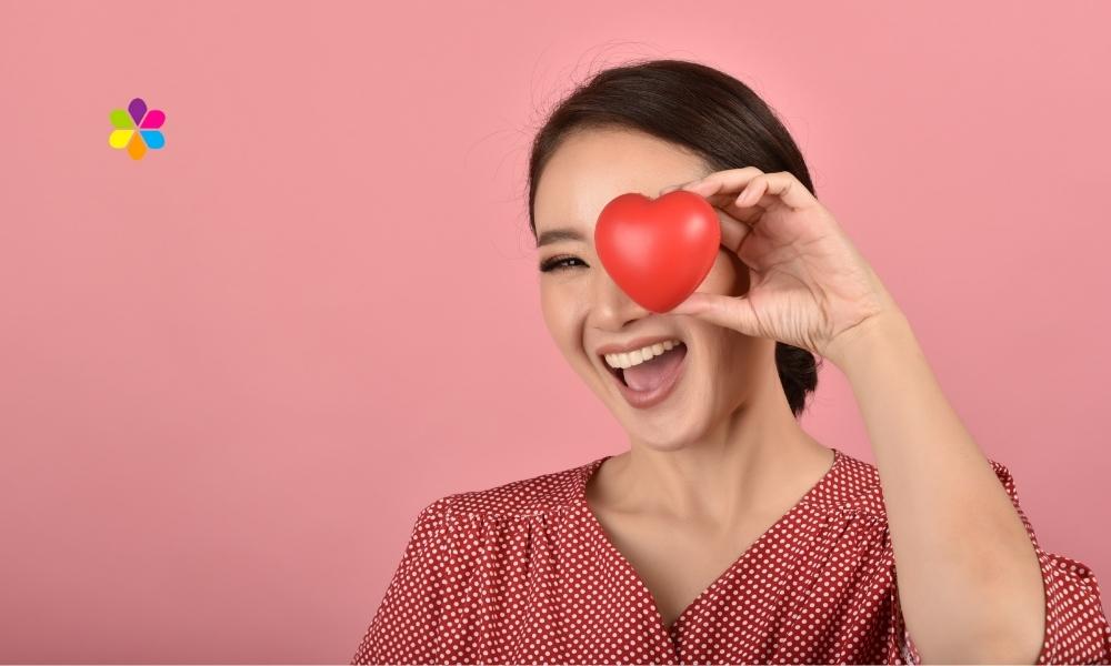 woman in red shirt holds red heart shape to her eye and smiles
