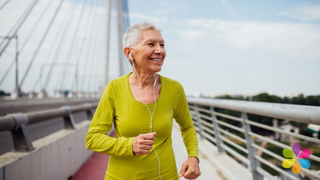 older smiling woman in green shirt walking along a bridge in the sunshine.