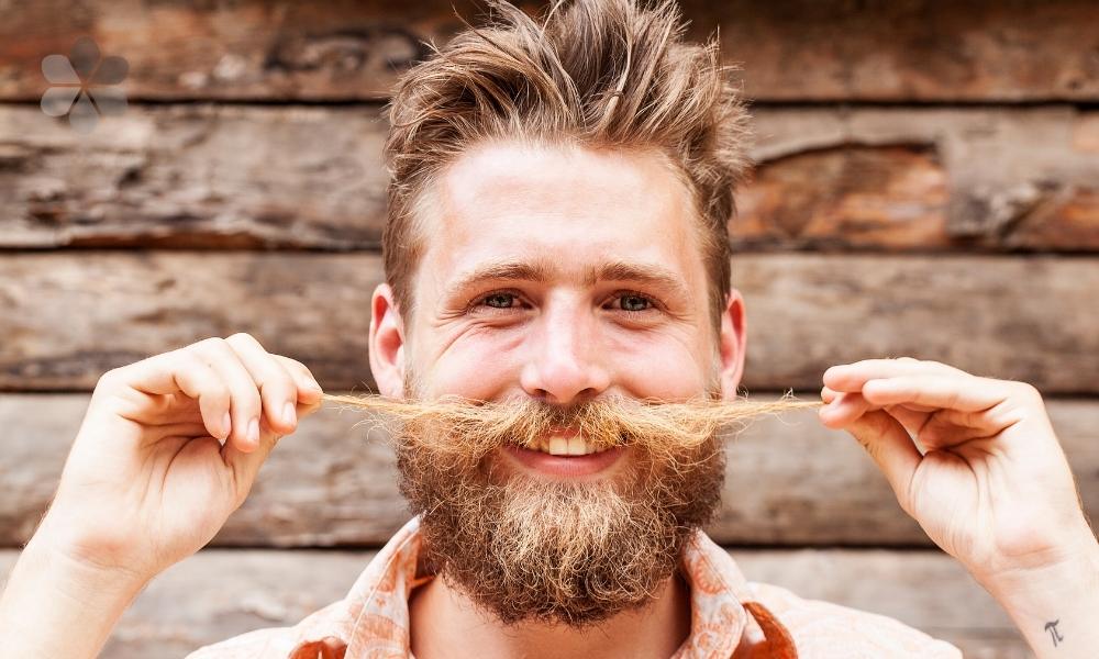 smiling fair-haired young man pulls both ends of his long moustache